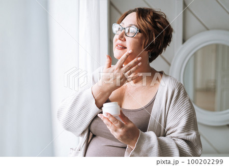Smiling middle aged plus size woman with day cream near window at home Smiling middle aged plus size woman with day cream near window at home 100202299