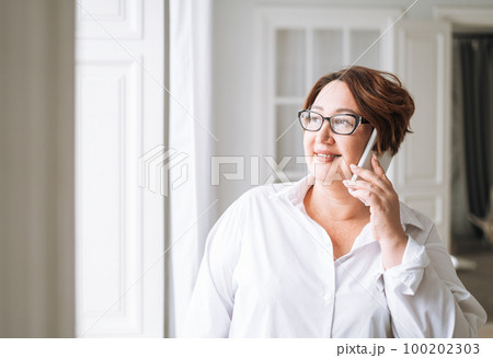 Adult charming brunette woman in glasses plus size body positive using mobile near window Adult charming brunette woman in glasses plus size body positive using mobile near window 100202303