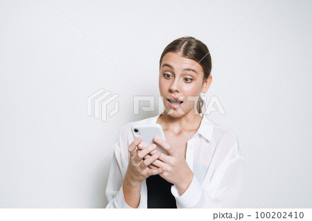 Surprised teenager girl with long hair in white shirt using mobile phone on white background Surprised teenager girl with long hair in white shirt using mobile phone on white background 100202410