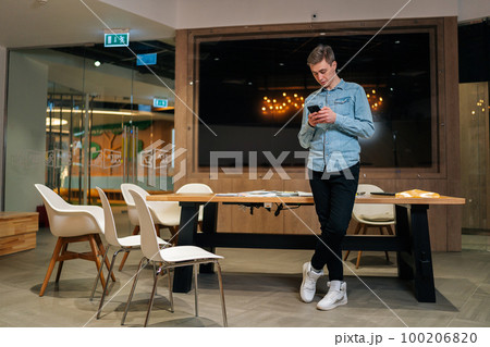 Portrait of handsome startup business man using online app on smartphone, standing by table at office boardroom with dark modern interior, reading text message, typing, chatting on internet. 100206820