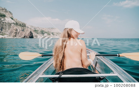 Woman in kayak back view. Happy young woman with long hair floating in transparent kayak on the crystal clear sea. Summer holiday vacation and cheerful female people relaxing having fun on the boat Woman in kayak back view. Happy young woman with long hair floating in transparent kayak on the crystal clear sea. Summer holiday vacation and cheerful female people relaxing having fun on the boat 100208088