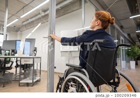 Red-haired caucasian woman in a wheelchair trying to open the door in the office. Red-haired caucasian woman in a wheelchair trying to open the door in the office. 100209346