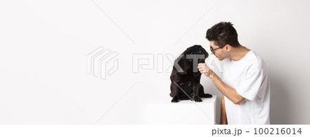 Handsome young hipster in glasses, pet his dog, looking at each other, standing over white background Handsome young hipster in glasses, pet his dog, looking at each other, standing over white background 100216014