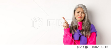 Judgy and skeptical asian senior woman grimacing displeased, pointing finger upper left corner unamused, standing over white background Judgy and skeptical asian senior woman grimacing displeased, pointing finger upper left corner unamused, standing over white background 100216016