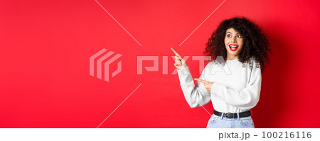 Excited pretty girl with curly hair and red lips, looking and pointing fingers left with amazed face, showing banner, standing on studio background Excited pretty girl with curly hair and red lips, looking and pointing fingers left with amazed face, showing banner, standing on studio background 100216116
