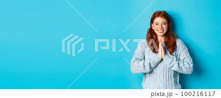 Cute redhead girl saying thank you, smiling and looking at camera, expressing gratitude, standing against blue background Cute redhead girl saying thank you, smiling and looking at camera, expressing gratitude, standing against blue background 100216117
