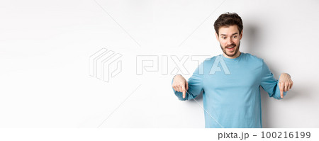 Handsome caucasian man pointing down with impressed and happy face, smiling and saying wow, standing on white background Handsome caucasian man pointing down with impressed and happy face, smiling and saying wow, standing on white background 100216199