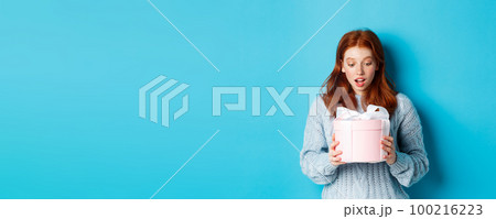 Beautiful redhead girl holding gift in pink cute box, looking at present with surprised face, standing over blue background Beautiful redhead girl holding gift in pink cute box, looking at present with surprised face, standing over blue background 100216223