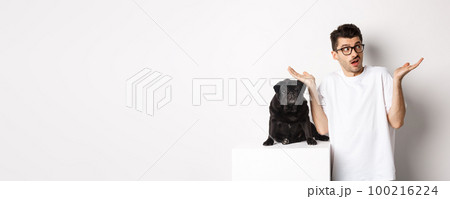 Portrait of confused hipster guy dog owner shrugging, standing near cute black pug pet, white background Portrait of confused hipster guy dog owner shrugging, standing near cute black pug pet, white background 100216224