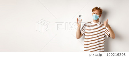 Covid-19, health and lifestyle concept. Smiling male model with red hair, wearing face mask, showing hand sanitizer and thumb-up, recommend to use antiseptic, white background 100216293