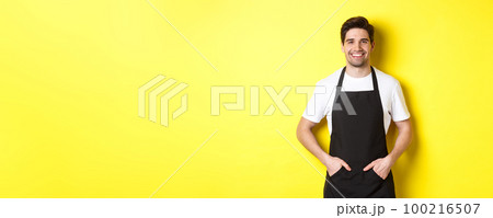 Happy barista in black apron looking at camera. Coffee shop owner wearing cafe uniform and smiling, standing over yellow background 100216507