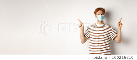 Covid, virus and social distancing concept. Handsome young man with red hair, wear face mask and pointing sideways at two promo offers, white background 100216810