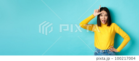 Asian girl winner mocking over lost team, showing loser sign on forehead and sticking tongue ignorant, standing over blue background Asian girl winner mocking over lost team, showing loser sign on forehead and sticking tongue ignorant, standing over blue background 100217004