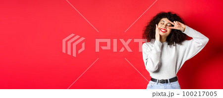 Beauty. Pretty lady with curly hair and red lips, touching face with makeup and showing v-sign on eye, smiling carefree, standing against studio background Beauty. Pretty lady with curly hair and red lips, touching face with makeup and showing v-sign on eye, smiling carefree, standing against studio background 100217005