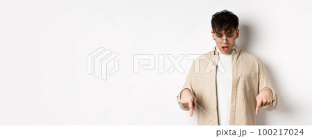 Amazed young guy in glasses look and point down, open mouth and say wow impressed, checking out discount offer, standing on white background 100217024