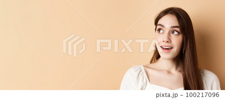 Dreamy beautiful girl looking at upper left corner logo, checking out banner, standing thoughtful on beige background 100217096