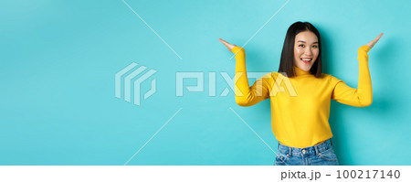 Beauty and fashion concept. Stylish beautiful asian girl demonstrate promotion, raising hands up and smiling, showing something on blue background Beauty and fashion concept. Stylish beautiful asian girl demonstrate promotion, raising hands up and smiling, showing something on blue background 100217140