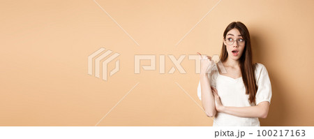 Excited girl in glasses pointing left, looking surprised at banner, standing on beige background 100217163