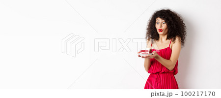 Excited birthday girl in red dress blowing candle on cake and making wish, standing on white background Excited birthday girl in red dress blowing candle on cake and making wish, standing on white background 100217170