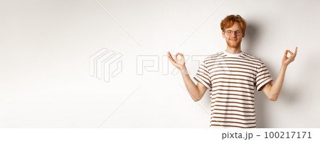 Calm and relaxed young man with red messy hair, spread hands sideways in mudra gesture and smiling, practice yoga or meditating, white background 100217171