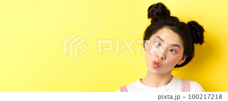 Close up portrait of funny asian teen girl with glamour makeup and hairstyle, squinting eyes and pucker lips silly, standing on yellow background 100217218
