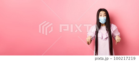 Healthy people and covid-19 pandemic concept. Excited asian girl in face mask pointing fingers down, showing advertisement with interest, standing against pink background 100217219