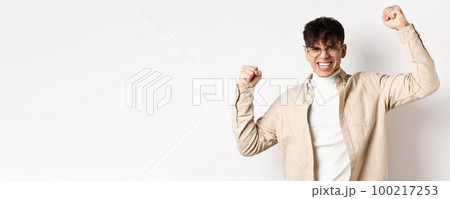 Portrait of motivated young man feeling joy, raising hands up and celebrating victory, triumphing and saying yes, standing on white background 100217253