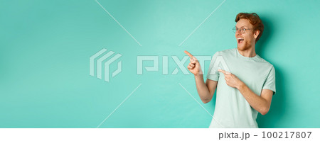 Excited young man with red hair, wearing glasses and t-shirt, pointing and looking left at awesome promotion, smiling happy at banner, turquoise background Excited young man with red hair, wearing glasses and t-shirt, pointing and looking left at awesome promotion, smiling happy at banner, turquoise background 100217807