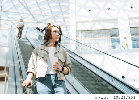 Young brunette teenager girl student in glasses on escalator in shopping mall public place 100217851