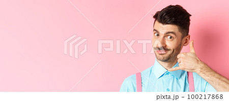 Close-up of handsome young guy with moustache, showing phone gesture, asking to call him, standing on pink background 100217868