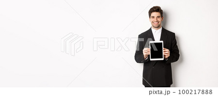Portrait of handsome businessman in trendy suit, showing digital tablet screen and smiling, standing against white background 100217888