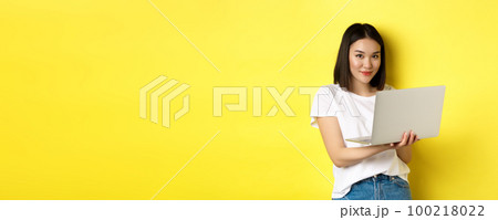 Young asian woman working freelance, using laptop and smiling, standing over yellow background Young asian woman working freelance, using laptop and smiling, standing over yellow background 100218022
