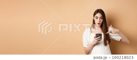 Online shopping concept. Excited girl pointing at smartphone screen and looking at camera amazed, standing against beige background Online shopping concept. Excited girl pointing at smartphone screen and looking at camera amazed, standing against beige background 100218023