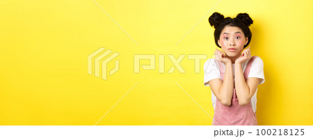 Cute teenage asian girl with beauty makeup, listen with interest, lean on hands and looking excited at camera, standing on yellow background 100218125