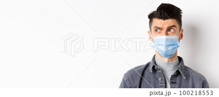 Pandemic lifestyle, healthcare and medicine concept. Confused guy in face mask look left at logo and frowning skeptical, standing on white background 100218553