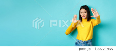 Image of woman feeling uncomfortable in front of camera, cover face with hands and frowning, begging to stop, standing against blue background 100221935