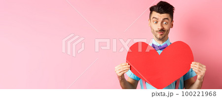 Valentines day concept. Cute boyfriend looking at big red heart, waiting for lover on romantic date, pink background 100221968