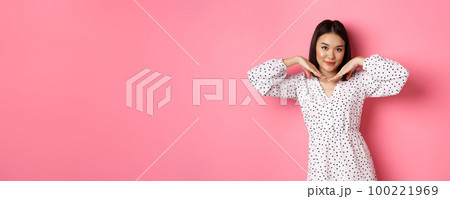Beautiful asian woman in trendy dress making cute face, holding hands near jawline and gazing coquettish at camera, standing over pink background 100221969