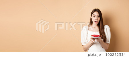 Excited birthday girl looking amazed, holding cake with candle, making b-day wish, standing on beige background 100222534