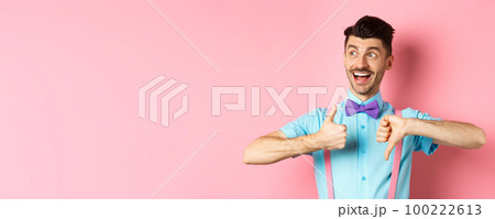 Cheerful man looking happy left, showing thumbs up down, judging product, give positive and negative feedback, standing over pink background 100222613