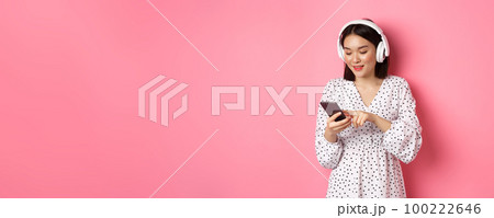 Beautiful asian woman texting message on smartphone, listening music in headphones, standing over pink background Beautiful asian woman texting message on smartphone, listening music in headphones, standing over pink background 100222646