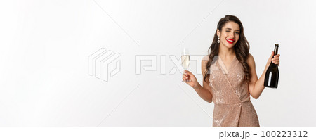 Winter holidays celebration concept. Beautiful woman enjoying Christmas and New Year party, drinking champagne and winking at camera, standing in elegant dress 100223312