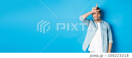 Rude guy mocking person for losing, showing loser sign on forehead and looking with dismay, standing against blue background 100223318