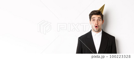 Close-up of attractive man in party hat and trendy suit, looking surprised, celebrating new year, standing against white background Close-up of attractive man in party hat and trendy suit, looking surprised, celebrating new year, standing against white background 100223328