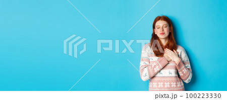 Image of dreamy redhead girl close eyes and holding hands on heart, feeling nostalgic, remember or imagine something, standing over blue background Image of dreamy redhead girl close eyes and holding hands on heart, feeling nostalgic, remember or imagine something, standing over blue background 100223330