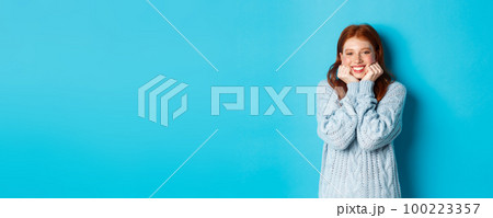 Lovely redhead teenage girl looking daydreaming, staring with admirationa sympathy at camera, standing against blue background 100223357