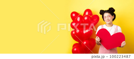 Happy Valentines day. Silly and beautiful asian woman smiling dreamy, showing red heart, imaging romantic date with lover, standing on yellow background 100223487