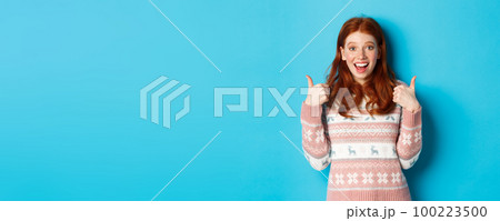 Girl approve something awesome, showing thumb-up, well done gesture and smiling amazed, standing over blue background 100223500