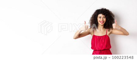 Attractive young woman recommending promo offer, showing thumb up and smiling, like product, standing in festive red dress on white background 100223548