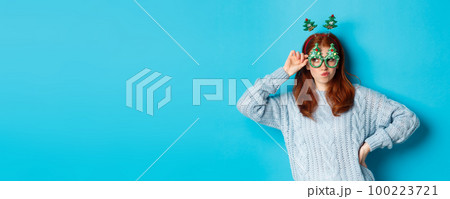 Winter holidays and Christmas sales concept. Beautiful redhead female model celebrating New Year, wearing funny party headband and glasses, smiling silly, blue background 100223721